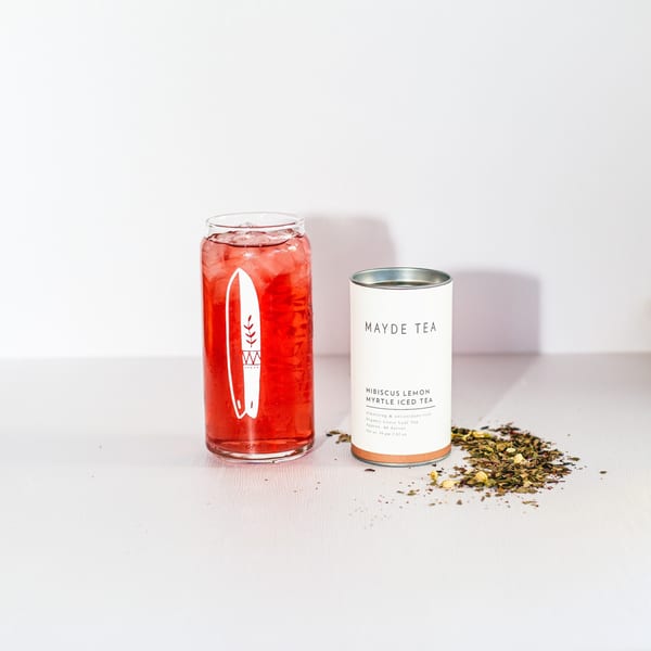 A glass of red iced tea with a surfboard logo next to a Mayde Tea container and loose tea.