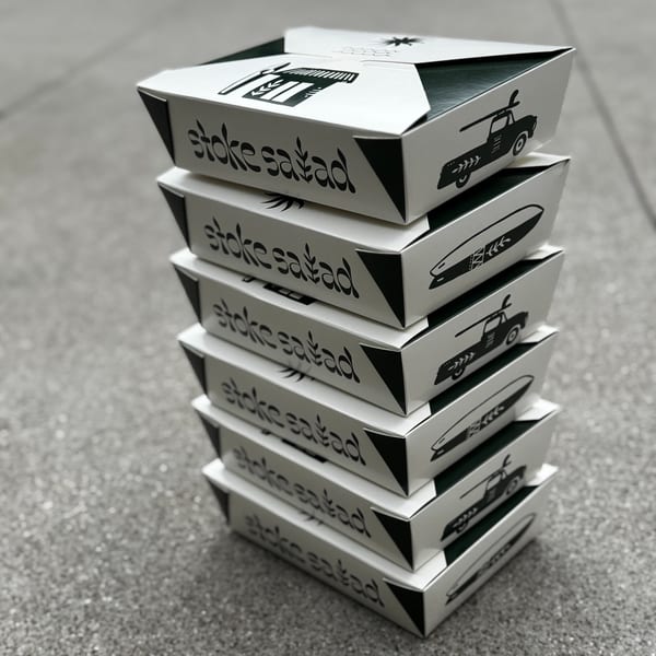 A stack of Stoke Salad takeout boxes with the brand logo and surf-themed illustrations.