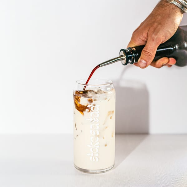 A hand pours dark liquid from a bottle into a Stoke Salad glass with a milky drink and ice.